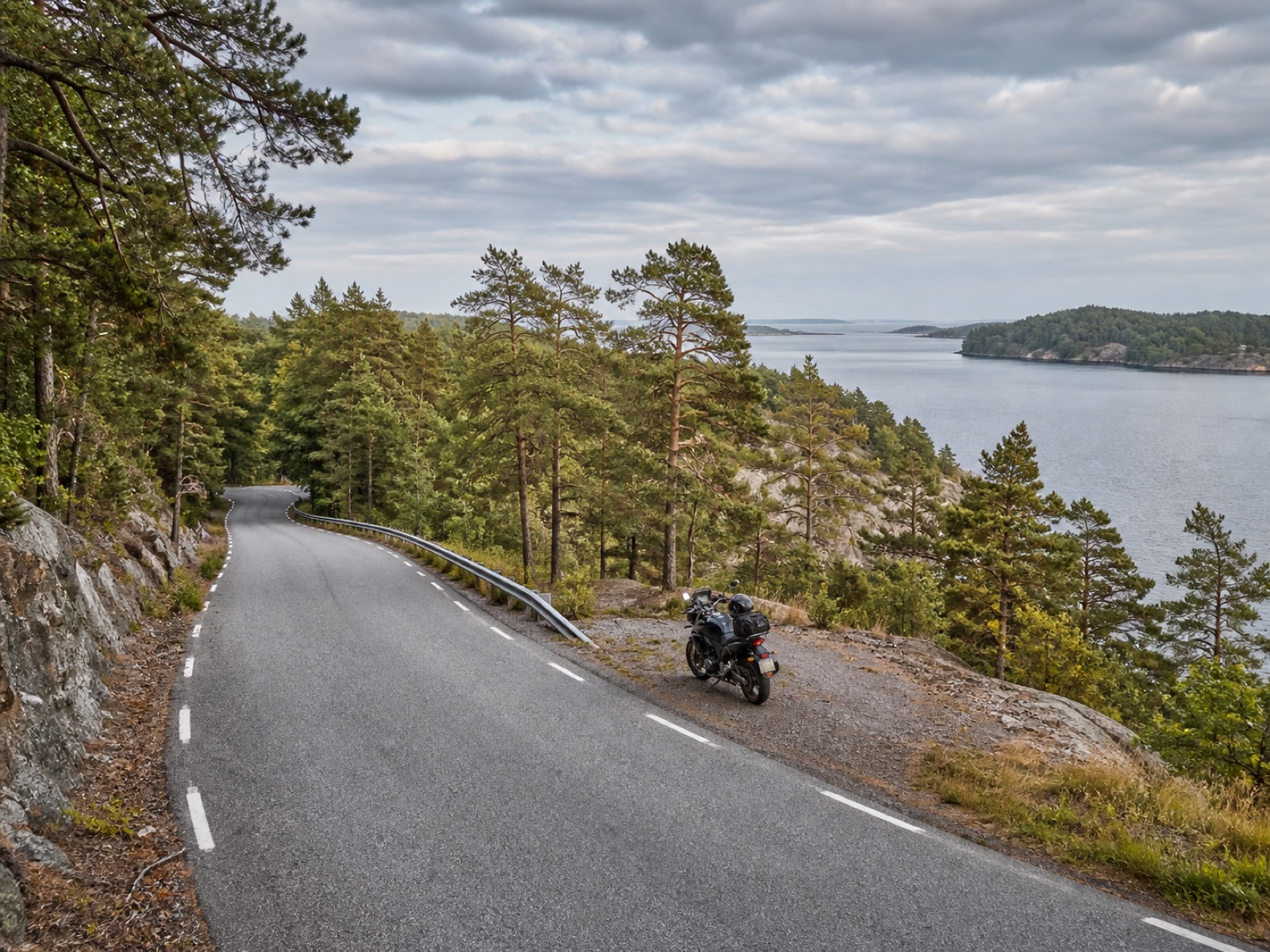 Motorcykel parkerad vid en slingrande kustväg i Roslagen med skog och vatten i bakgrunden